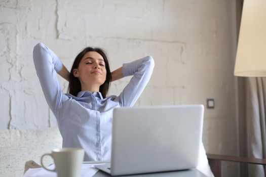 Young woman crossed hands behind head, enjoying break time at home. Peaceful carefree business woman resting at table with computer, looking aside, dreaming of future