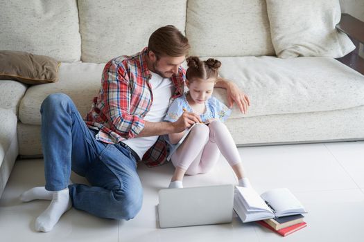 Smiling father and daughter sitting on floor in living room with laptop, teaching lessons