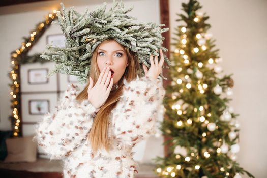 Surprised woman opening eyes wide and holding hand to her mouth while wearing a crown made of fir