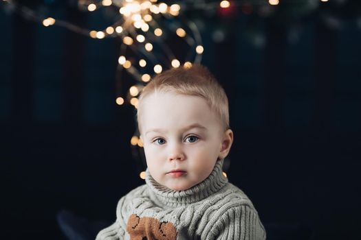 Close-up photo of a serious little kid looking at the camera with Christmas lights in background