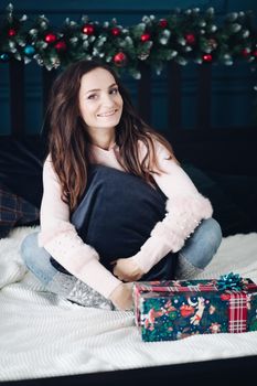 Lovely brunette woman sitting cross-legged on a bed near a wrapped gift and smiling