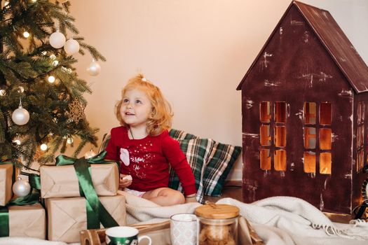 Cute blonde child with marshmallow in hand sitting under decorated Christmas tree and admiring it.