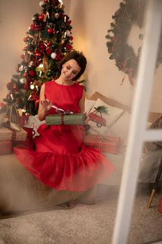 Portrait of a beautiful brown-haired woman in red dress sitting on bed and opening Christmas gift.