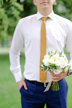 Young european groom keeping bouquet of flowers and wearing white shirt. Concept of wedding photo session and waiting for bride.