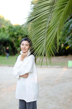 Korean pretty woman in white sweater standing near palm leaf. Concept of asian beauty and female person on tropical resort.