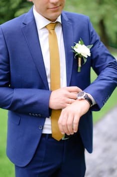 Young groom looking at watch and wearing blue jacket with flower. Concept of wedding photo session and waiting for bride.