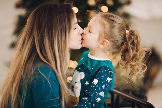 Close-up photo of a brunette lady kissing a little girl with Christmas tree in the background. Holiday concept