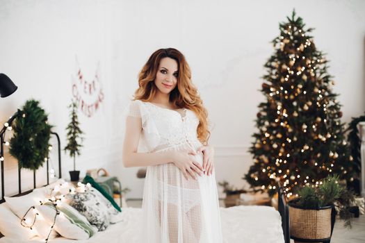 Handsome pregnant caucasian woman poses for the camera in white dress in christmas atmosphere