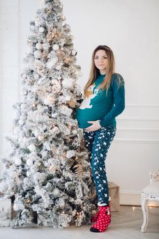 Attractive pregnant woman in sleepwear poses near the christmas tree aand smiles