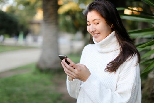 Chinese smiling girl using smartphone and walking in tropical park. Concept of modern technology and nature.