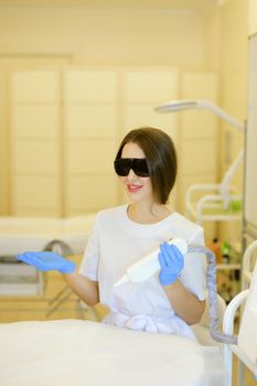 Young female cosmetologist wearing special purpose glasses and latex gloves sitting near permanent makeup device. Concept of beauty cosmetology salon and expensive high quality equipment.