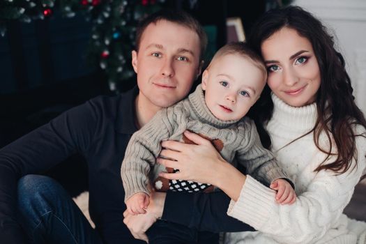 Beautiful mom, young dad and their little baby sit next to each other on Christmas Eve