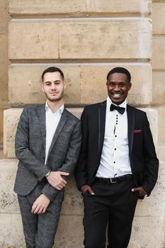 Afro american and caucasian smiling gays standing near building and wearing suits. Concept of lgbt and walking in city.