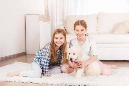 Smiling sisters sit together on carpet with cute retriever puppy