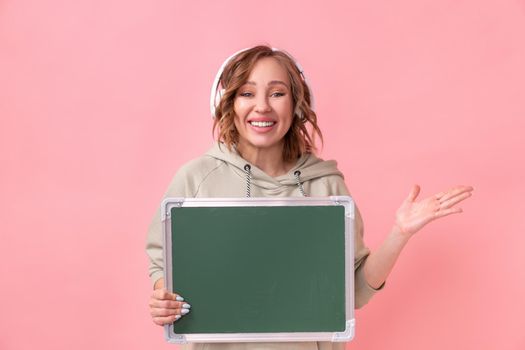 Woman holding empty chalkboard over pink background Happy Caucasian female 30 years old dressed headphones oversize hoodie hold blank green board sign Studio shoot Advertising Promoting Presentation