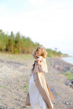 Young woman wearing coat and white dress standing on sea beach with trees in background. Concept of summer vacations photo session and freedom, happiness.