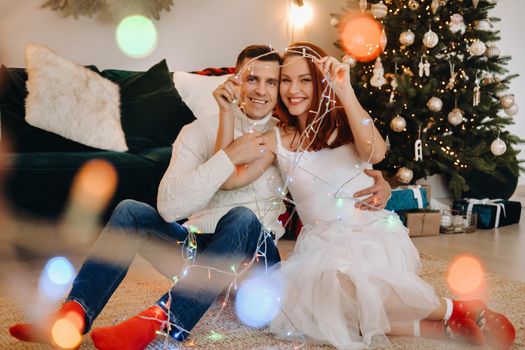 a happy married couple is sitting at home on the floor near the Christmas tree.