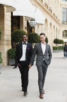 Afro american and caucasian happy handsome gays walking outside and holding hands in city. Concept of same sex male couple.