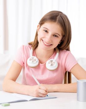 Pretty school girl making homework with listening to online lessons on headphones