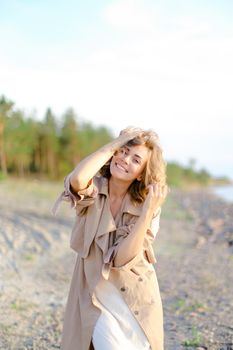 Young caucasian girl wearing coat and white dress standing on sea beach with trees in background. Concept of summer vacations photo session and freedom, happiness.