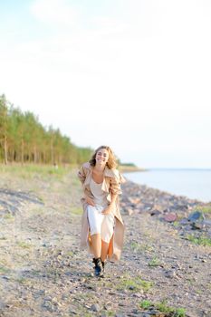 Smiling blonde woman wearing coat and white dress standing on sand with trees in background. Concept of happiness, youth and fashion, summer vacations.