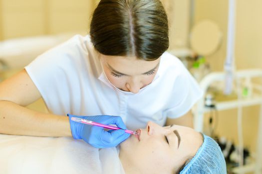 Skilled cosmetologist shaping lips with pencil for young girl before microblading. Concept of permanent makeup artist and beauty salon.