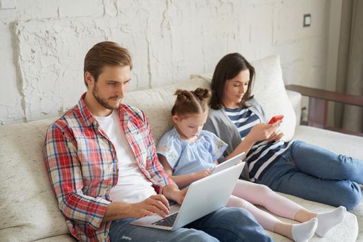 Father, mother and daughter using electronic devices sitting on sofa at living room