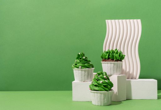 Merry christmas and happy new year. Composition of green Christmas tree shaped cupcakes on white poduims, abstract and modern minimal style, green solid background, copy space