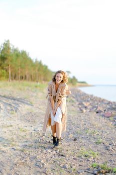 Blonde young woman standing on sea beach and wearing coat with white dress. Concept of happiness, youth and fashion, summer vacations.