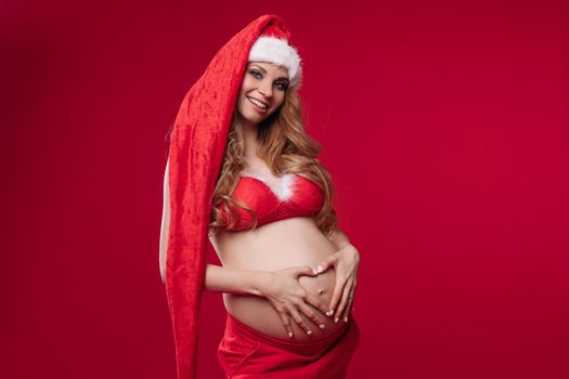 Charmilng young pregnant female in christmas costume smiles, picture isolated on red background