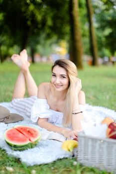 Young blonde pretty girl lying on plaid in park, reading book near watermelon and hat. Concept of leisure time, summer vacations and picnic.