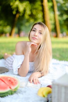 Young caucasian blonde woman lying on plaid near watermelon in park and reading book. Concept of picnic on nature, summer vacations and leisure time.