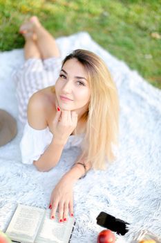 Young european girl with red nails lying on plaid in park and reading book. Concept of having free time, resring and picnic.