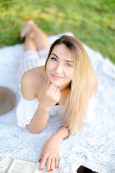 Young blonde pretty girl with red nails lying on plaid in park and reading book. Concept of having free time, resring and picnic.