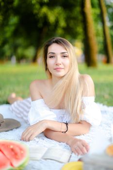 Young nice woman lying on plaid near watermelon in park and reading book. Concept of picnic on nature, summer vacations and leisure time.