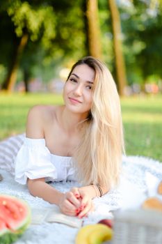 Young european woman lying on plaid near watermelon in park and reading book. Concept of picnic on nature, summer vacations and leisure time.