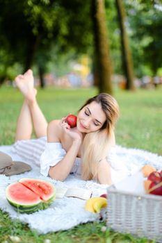 Young blonde pretty girl lying on plaid in park, keeping apple and reading book near watermelon and hat. Concept of leisure time, summer vacations and picnic, healthy food.