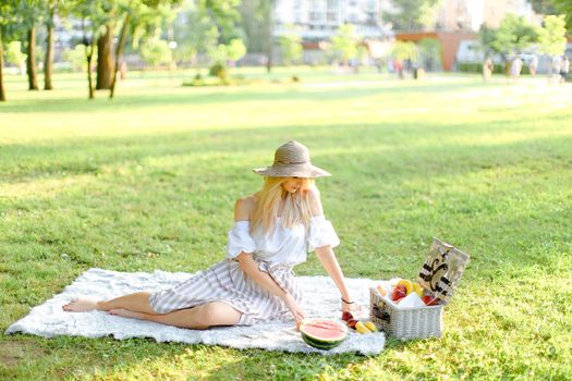 Young beautiful gitrl sitting in park on plaid near fruits, grass in background. Concept of summer picnic, resting on nature and healthy food.