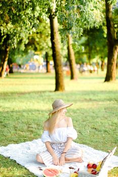 Young caucasian woman in hat sitting in park on plaid near fruits, grass and trees in background. Concept of summer picnic, resting on nature and healthy food.