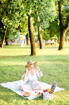 Young european woman in hat sitting in park on plaid near fruits, grass and trees in background. Concept of summer picnic, resting on nature and healthy food.