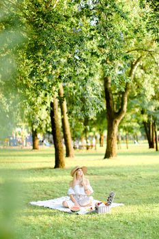 Young caucasian blonde woman in hat resting on plaid with fruits in garden. Concept of summer picnic in park, nature and free time.