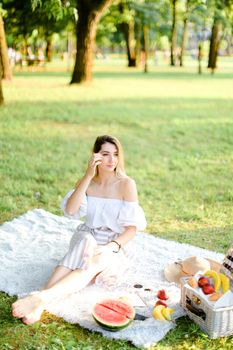 Young caucasian girl having picnic on plaid and sitting in park with fruits. Concept of resting in open air, leisure time and summer season