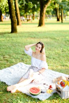 Young girl sitting on plaid with fruits in park. Concept of summer picnic, photo session in open air and vacations.