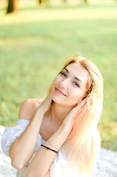 Portrait of young blonde happy woman sitting on plaid, grass in background. Concept of beautiful female person and summer season.