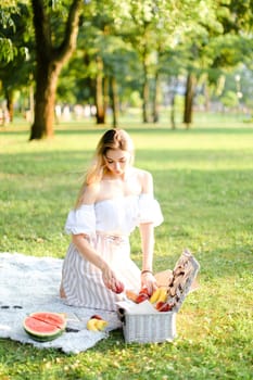 Young blonde woman preparing for picnic in park, sitting on plaid near box and waterlemon. Concept of having leisure time, resting in open air and summer season.