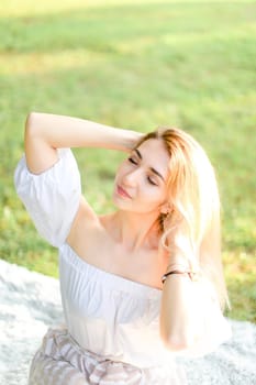 Portrait of young blonde caucasian woman sitting on plaid, grass in background. Concept of beautiful female person and summer season.