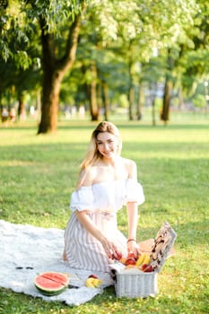 Young happy woman preparing for picnic in park, sitting on plaid near box and waterlemon. Concept of having leisure time, resting in open air and summer season.