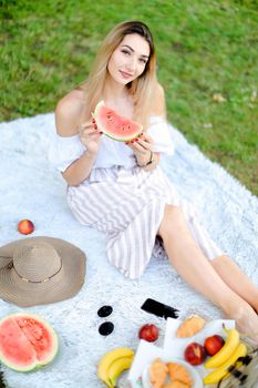 Young nice girl sitting on plaid near fruits and hat, eating watermelon, grass in background. Concept of summer picnic and resting on weekends in open air.