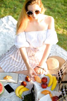 Young woman in sunglasses having picninc and sitting on plaid near opened book, fruits and smartphone. Concept of vegeterian lifestyle, leisure time and rest.