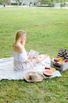 Young girl reading book and sitting on plaid near fruits in park. Concept of having summer picnic and leisure time.